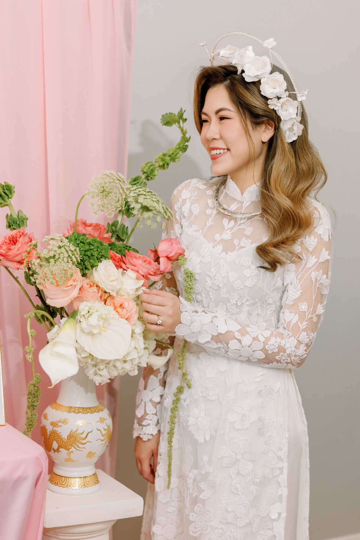 Bride in floral lace dress, smiling while arranging flowers in white and gold vase, pink curtain in background.