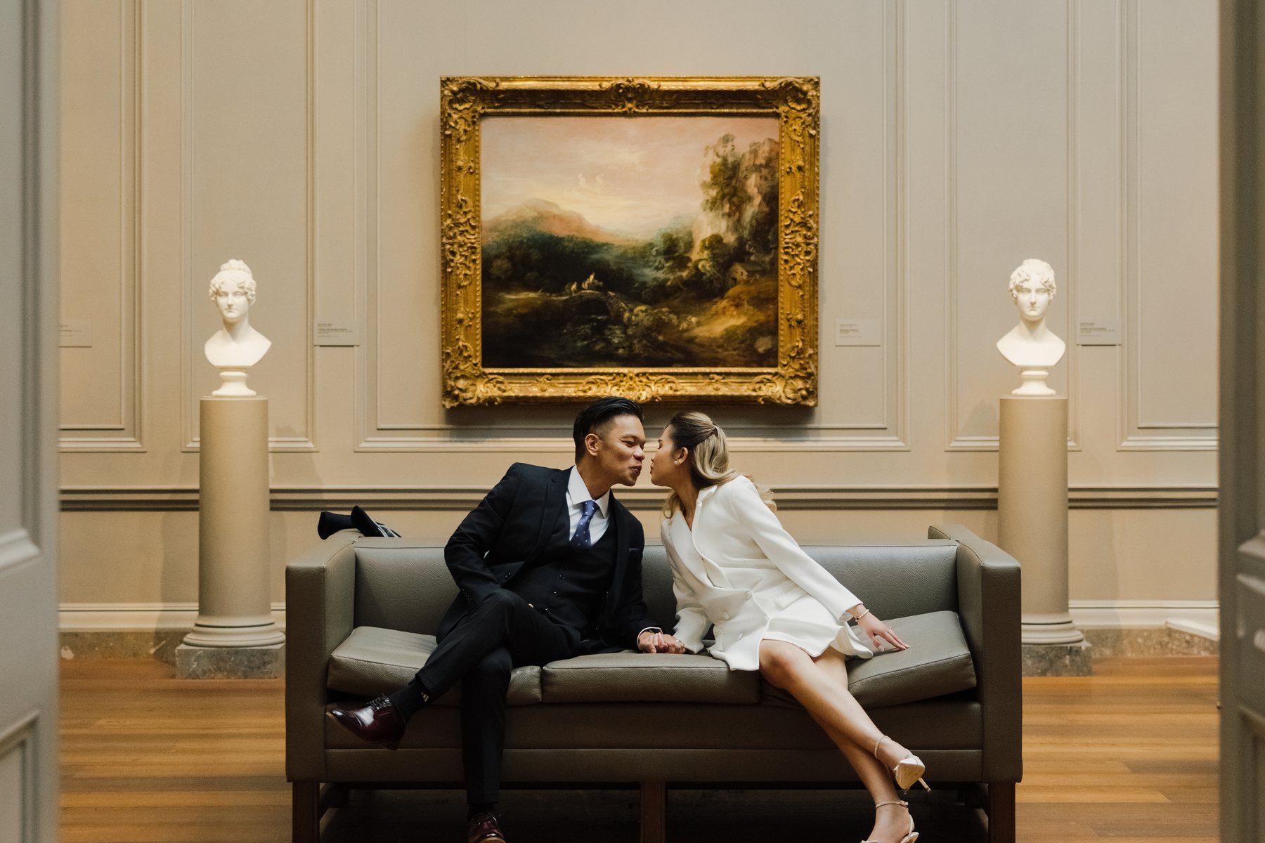 Engaged couple sharing a kiss on a gallery bench beneath a landscape painting at the National Gallery of Art, Washington, DC — photographed by Kevin Socola Films