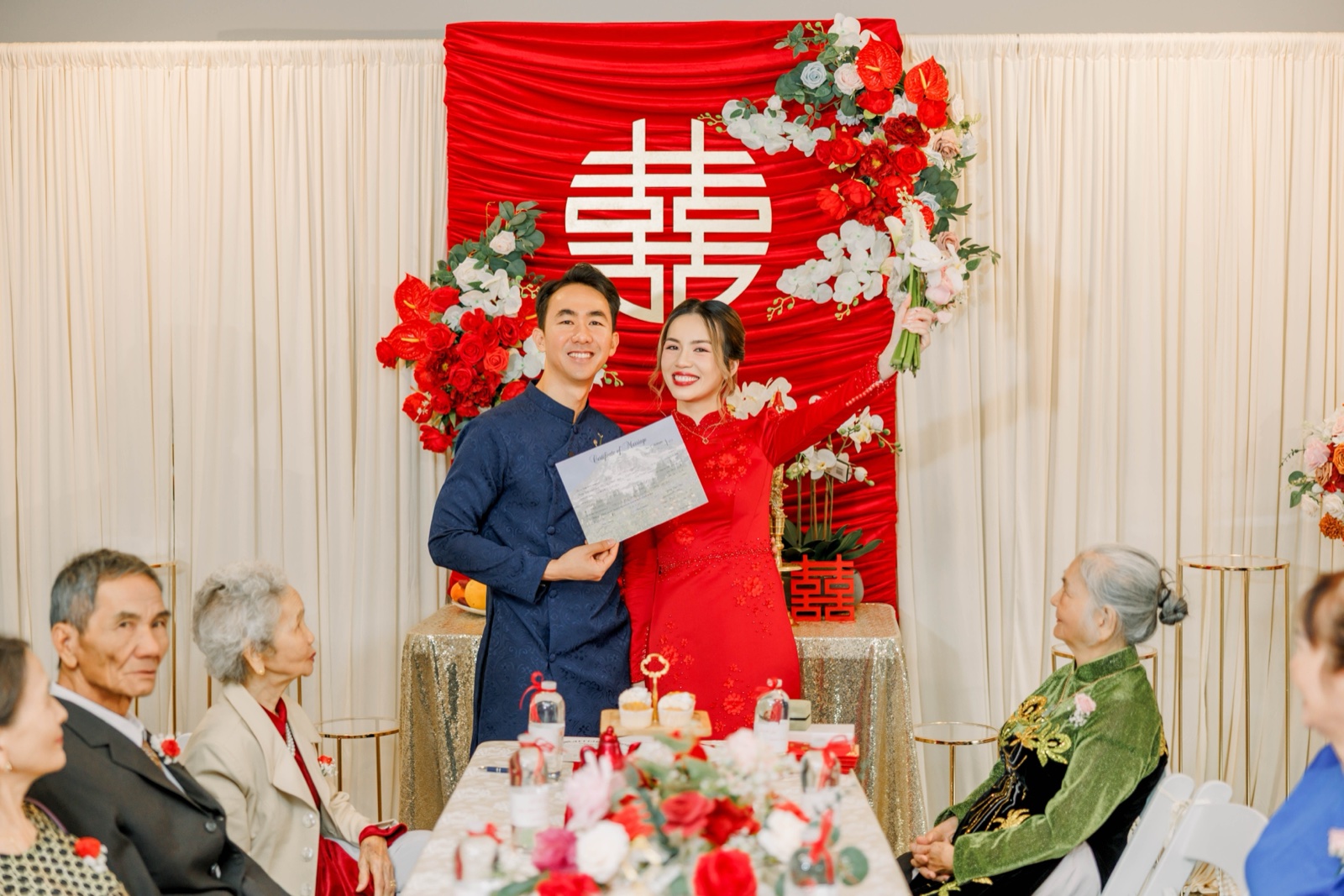 Vietnamese tea ceremony wedding — bride in red áo dài and groom in navy áo dài with double happiness 囍 backdrop and elders watching, photographed by Kevin Socola Films in the DMV