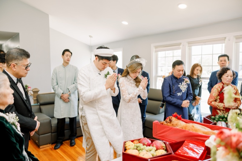 Vietnamese lễ gia tiên ancestor ceremony — bride and groom in white áo dài with traditional khăn đóng headpiece bowing in prayer before family altar with mâm quả and roasted pig offering, photographed by Kevin Socola Films in the DMV